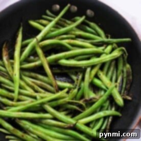 Green beans are sizzling in a black frying pan on a white surface, perfectly capturing the essence of how long to cook fresh green beans on the stove for that ideal flavor and crispness.