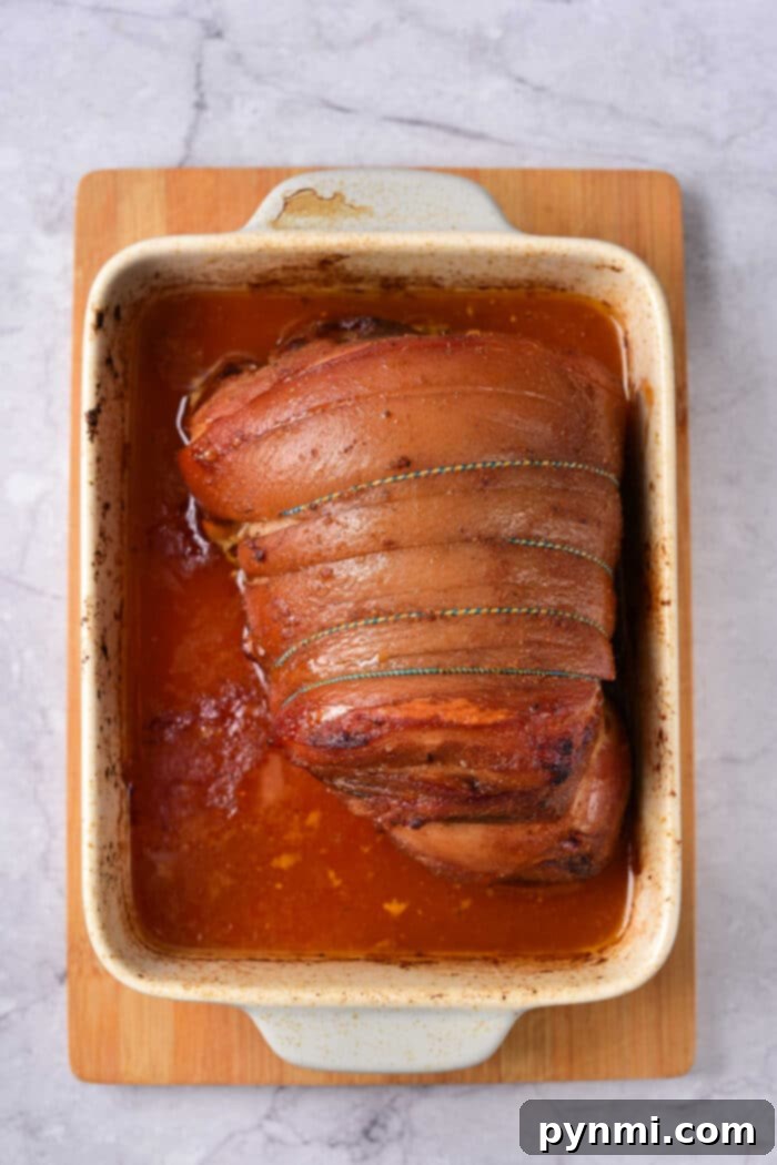 Mastering Pork Shoulder: How Long at 350°F for Ideal Tenderness 5 Roast pork shoulder with tied skin sits in a pan of reddish-brown sauce on a wooden board, perfect for those wondering how long to cook pork shoulder at 350 for tender, flavorful results.
