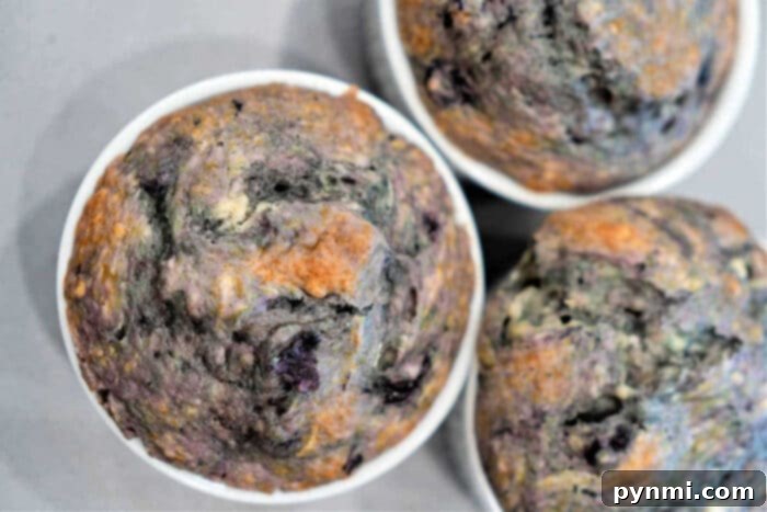 A close-up image of three Cake Mix Blueberry Muffins. The muffins have a golden-brown crust with visible blue and purple specks from the blueberries. They are in white paper muffin cups and are placed on a light-colored surface.