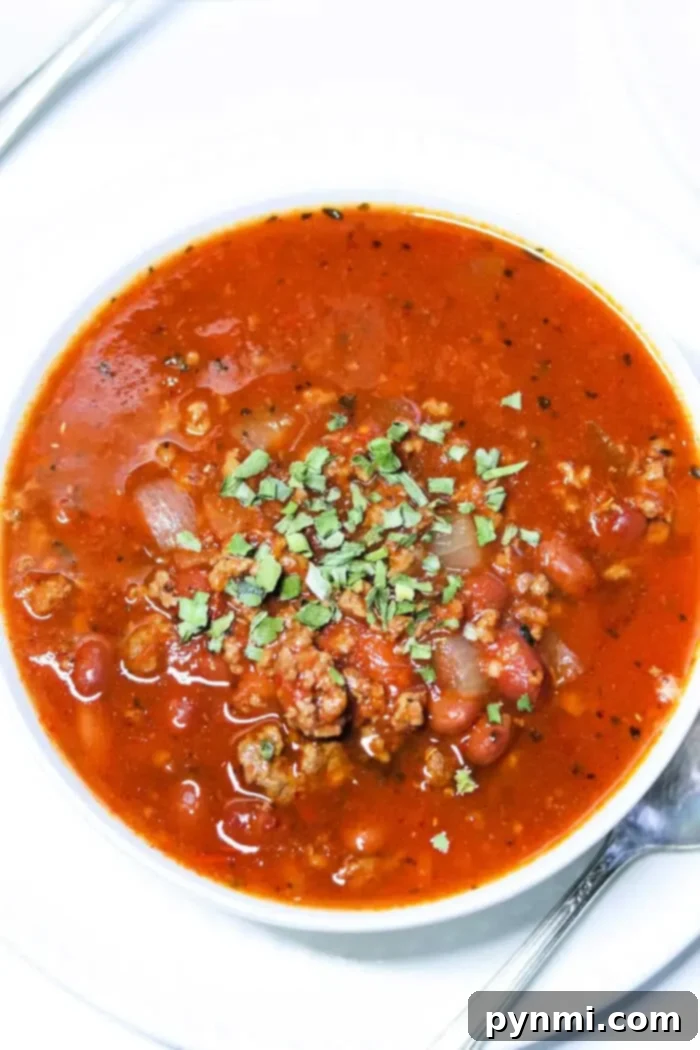 A bowl of chunky bear meat chili topped with chopped herbs. The hearty mix features beans, onions, and tender ground meat, all served in a pristine white bowl with a spoon ready on the side.