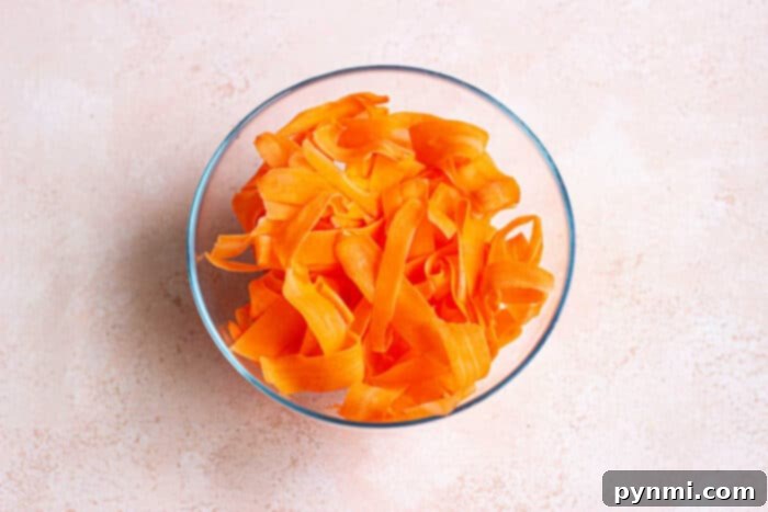 A glass bowl brimming with raw carrot salad featuring vibrant orange ribbons, set against a light beige background.