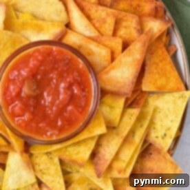 A plate of yellow and orange tortilla chips, made using an air fryer for a crispy texture, surrounds a bowl of red salsa. The plate rests on a surface where a green cloth peeks out from underneath.