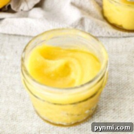 A small glass jar filled with smooth yellow lemon curd rests invitingly on a textured beige surface, enticing thoughts of delectable lemon curd desserts.