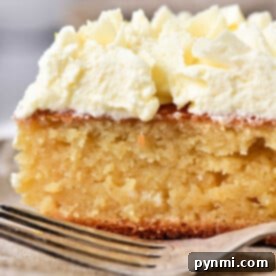 Close-up of a slice of moist lemon curd cake topped with whipped cream, placed on a plate with a fork nearby.