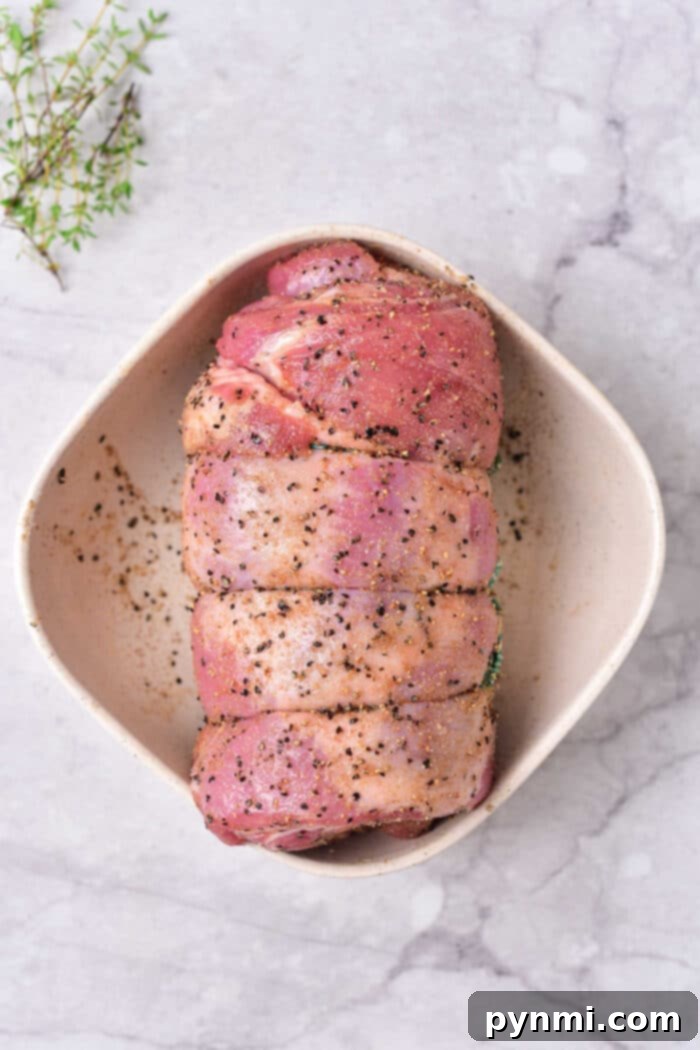 Succulent Lamb Breast Secrets 3 Raw seasoned rolled lamb breast in a white dish, primed for cooking, rests on a marble surface with a sprig of thyme nearby—a scene reminiscent of an exquisite lamb breast recipe.