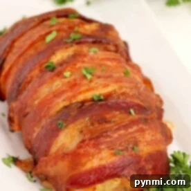 Meatloaf wrapped in crispy bacon, garnished with parsley, is elegantly served on a white plate with fresh herbs in the background.