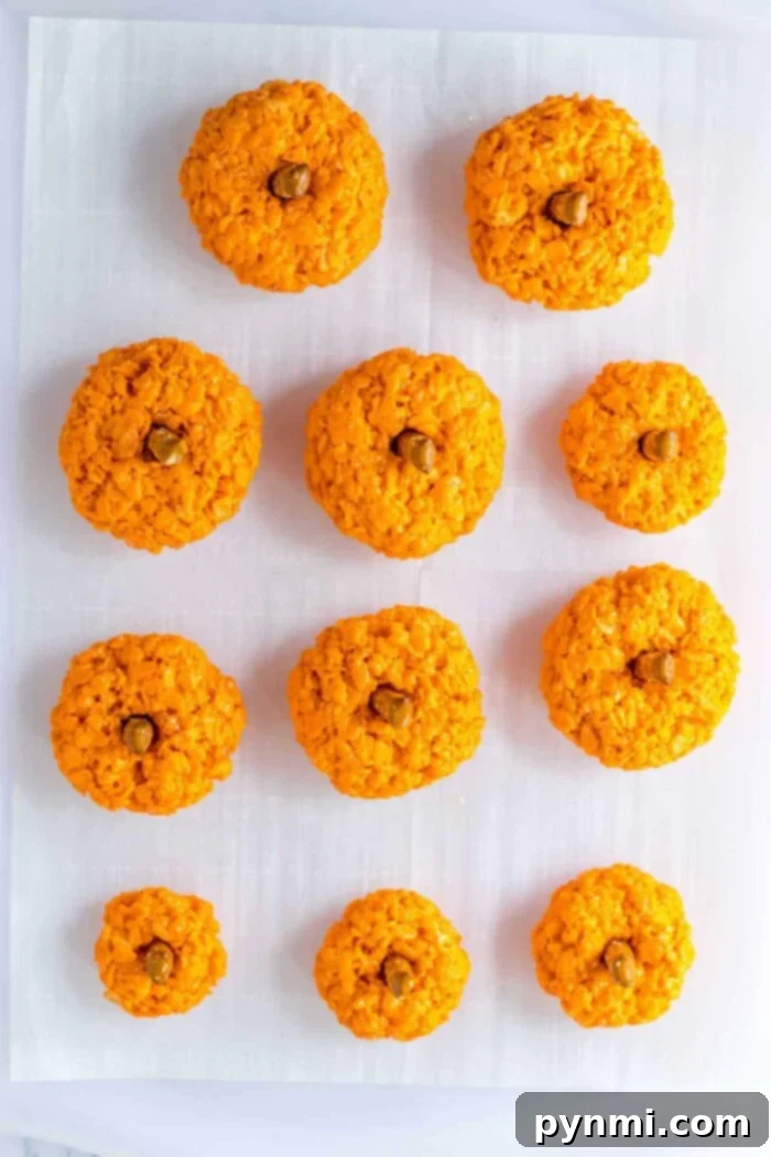 Ten Halloween Rice Krispie Treats shaped like pumpkins with green stems are arranged in rows on white parchment paper.