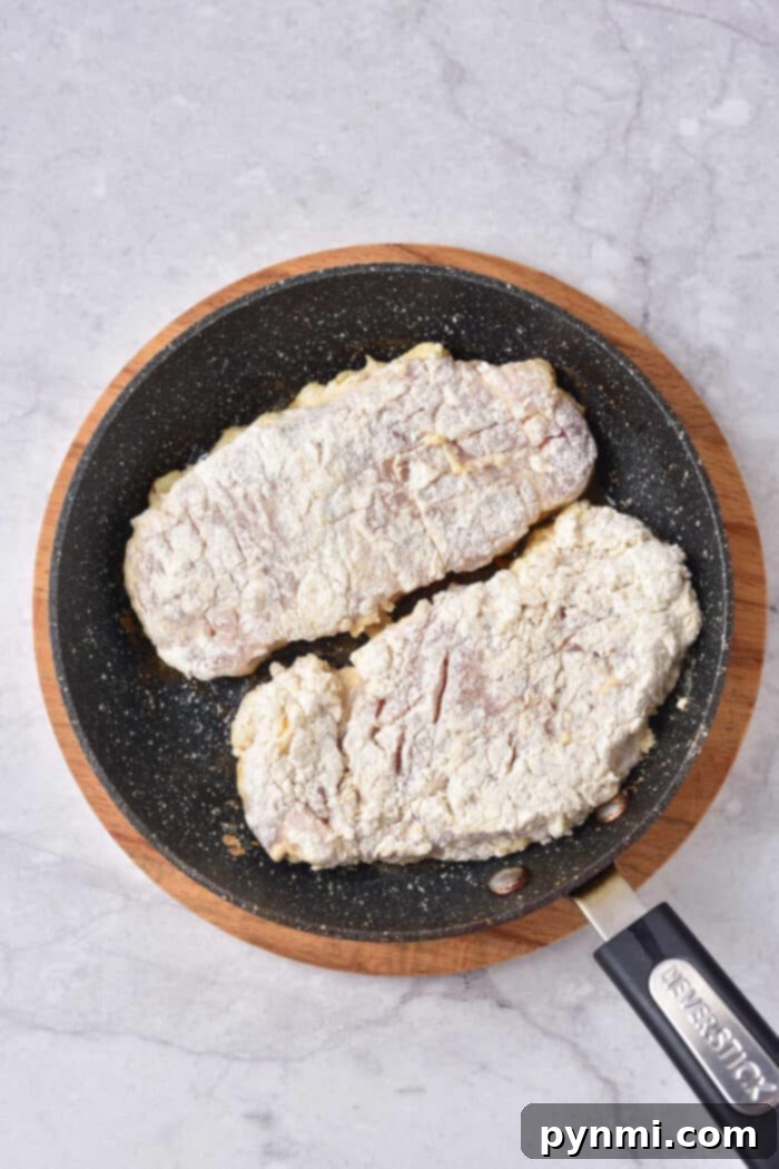 Tenderized Pork Cutlets 4 Two flour-coated pork steaks sizzle alongside a perfectly seasoned pork cube steak in a black frying pan on a marble surface.