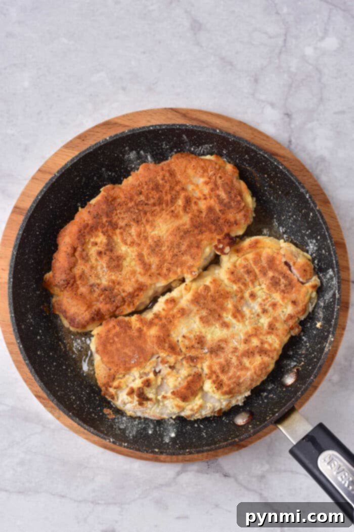 Tenderized Pork Cutlets 5 Two breaded pork cutlets and a succulent pork cube steak sizzle in a black pan set on a wooden board.
