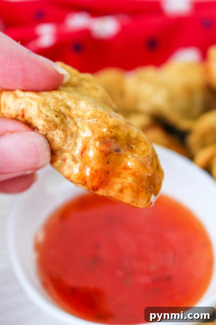 Close-up of a hand holding a golden-brown tempura chicken nugget, dipped in a bowl of red sauce. A red cloth adds warmth to the background.