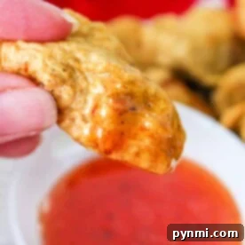 Close-up of a hand holding a golden-brown tempura chicken nugget, dipped in a bowl of red sauce. A red cloth adds warmth to the background.