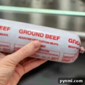 Hand holding a tube of ground beef in front of a stainless steel oven, ready to make delicious Ground Beef in Oven recipes.