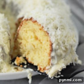 A slice of moist white chocolate coconut bundt cake with creamy white frosting and shredded coconut on a spatula, ready to be served.