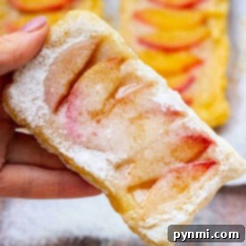 A hand delicately holds a flaky peach puff pastry dusted with powdered sugar, with more of these tempting pastries in the background.