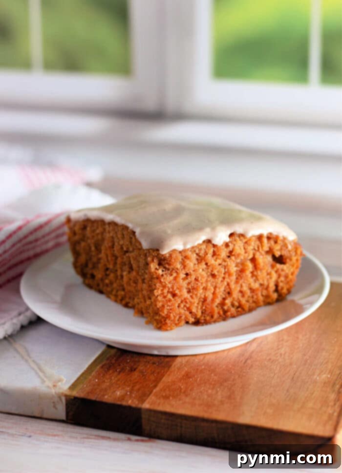 Golden Pumpkin Delight 5 A slice of frosted Crazy Cake carrot cake sits on a white plate. The cake has a thick, soft texture and is topped with a light layer of white icing. The plate rests on a wooden board, with a white and red striped cloth partially visible in the background.
