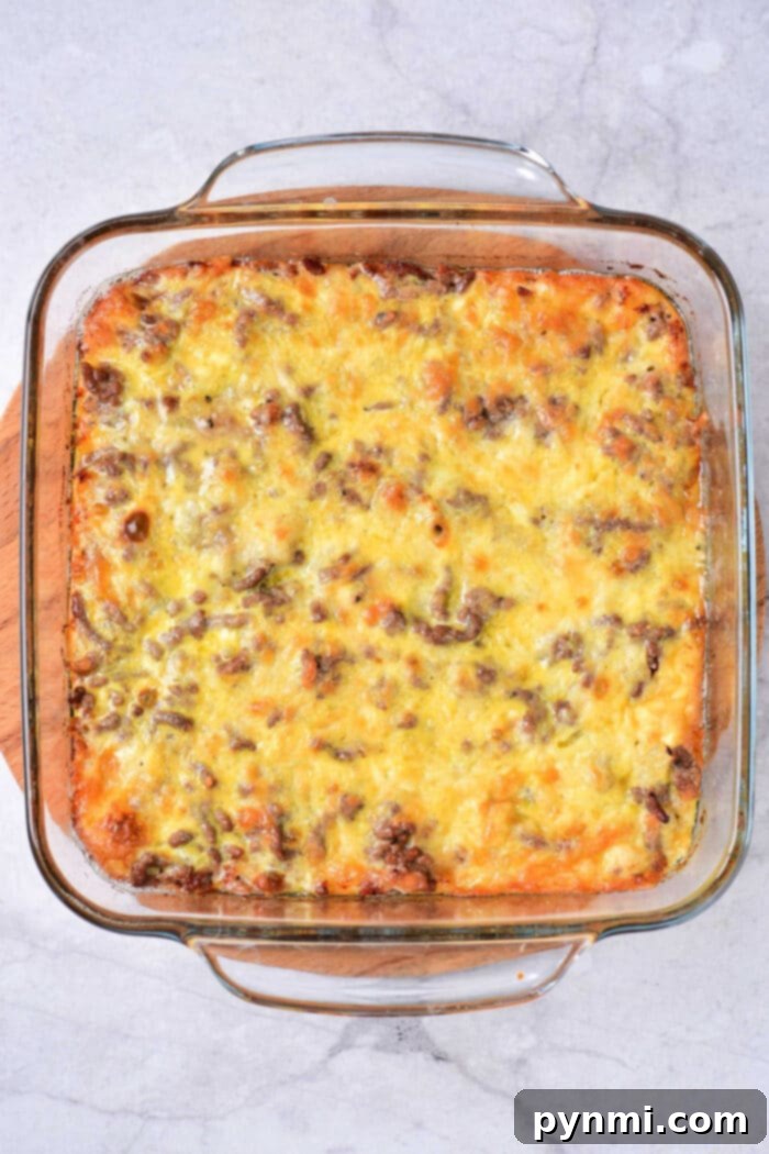 Ultimate Big Mac Bake 4 A Big Mac Casserole with a golden-brown cheese topping sits in a clear glass baking dish. The casserole, featuring ground meat and layers of melty cheese, boasts a slightly crispy surface. It rests invitingly on a white marble countertop, promising classic flavors in every bite.