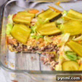 Glass dish with a layered Big Mac Casserole, topped with sliced pickles and crisp lettuce, one mouthwatering portion removed.