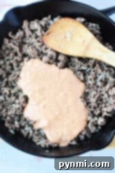 Ultimate Big Mac Bake 8 Ground beef simmered in a creamy, Big Mac-inspired sauce, mingling with sautéed onions in a skillet. A wooden spatula rests nearby, ready to serve these delightful Sloppy Joes.