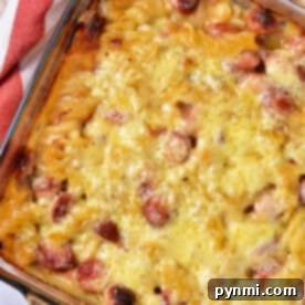 A baked dish in a glass pan, this hot dog casserole boasts a golden crust with visible slices of sausages and pasta, possibly macaroni. A red and white cloth is partially visible on the side.