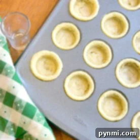 Mini cookie cups in a mini muffin pan beside a green checkered oven mitt and a small glass on the wooden surface.