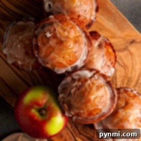 Freshly baked apple fritter muffins with glaze on a wooden board, complemented by two green and red apples, evoke the essence of baked apple fritters.