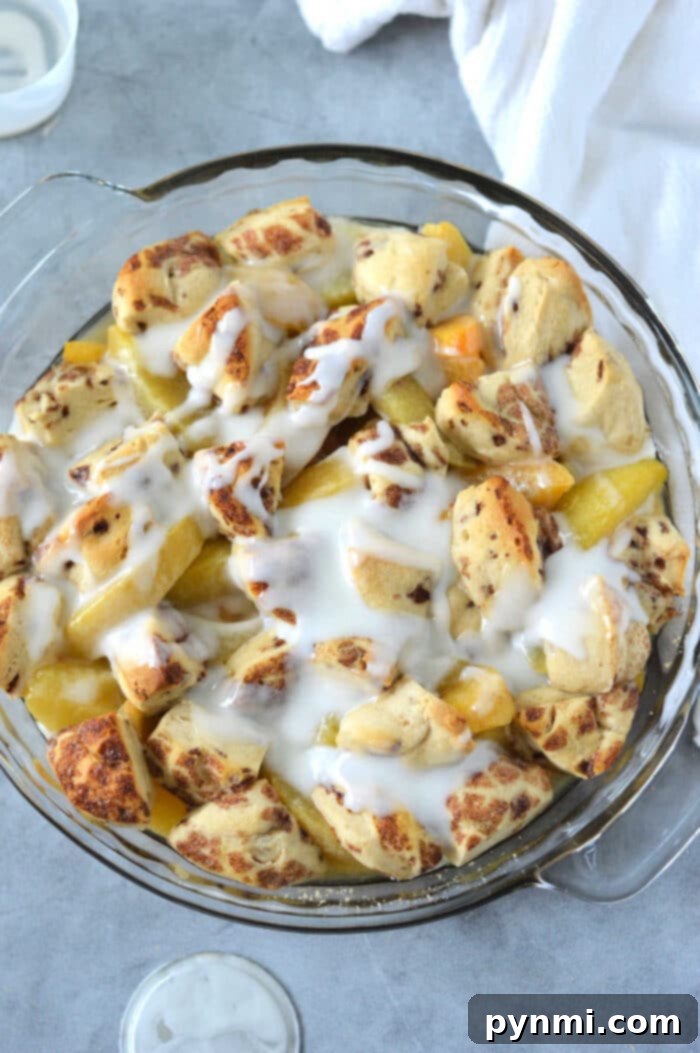 Peachy Cinnamon Swirl Cobbler 7 A glass dish filled with cinnamon roll biscuit pieces, peaches, and a drizzle of white icing evokes the comforting flavors of a cinnamon roll peach cobbler on a light gray surface.