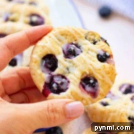 Fluffy Blueberry Biscuits 8 A person holding an easy biscuit with blueberries, drizzled with icing; more pastries on a plate in the background.