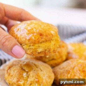 A hand holding a glazed honey biscuit delicately over a plate of assorted biscuits.