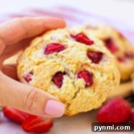 A hand holding a strawberry biscuit with visible pieces of the luscious fruit, reminiscent of a delightful strawberry biscuit, against a light background.