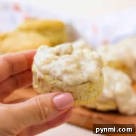 A hand holds a biscuit topped with creamy white vegan gravy, with more vegan biscuits in the background.