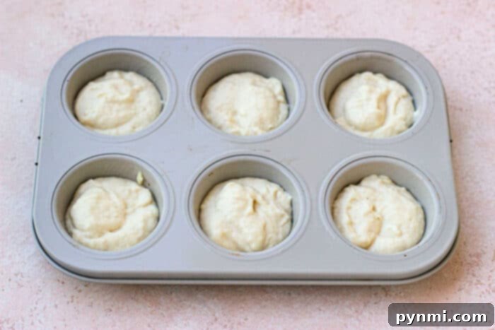 A muffin tin filled with six portions of creamy, unbaked batter sits gracefully on a light pink surface, ready to bake into mayonnaise biscuits.