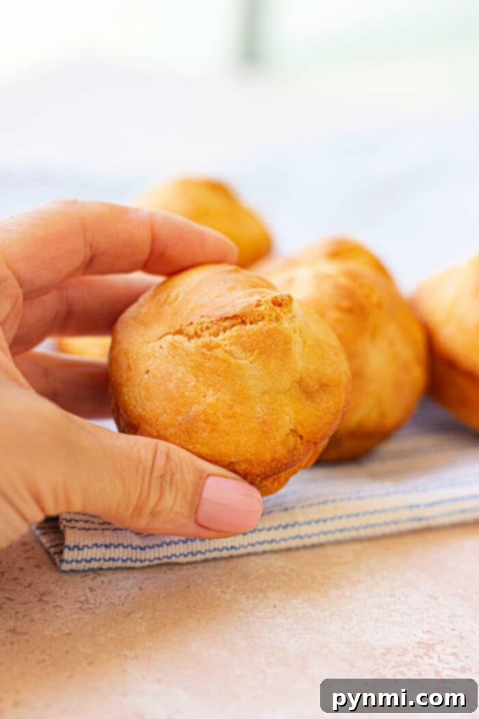 A hand holding a freshly baked mayo biscuit over a striped cloth, with more muffins tempting in the background.