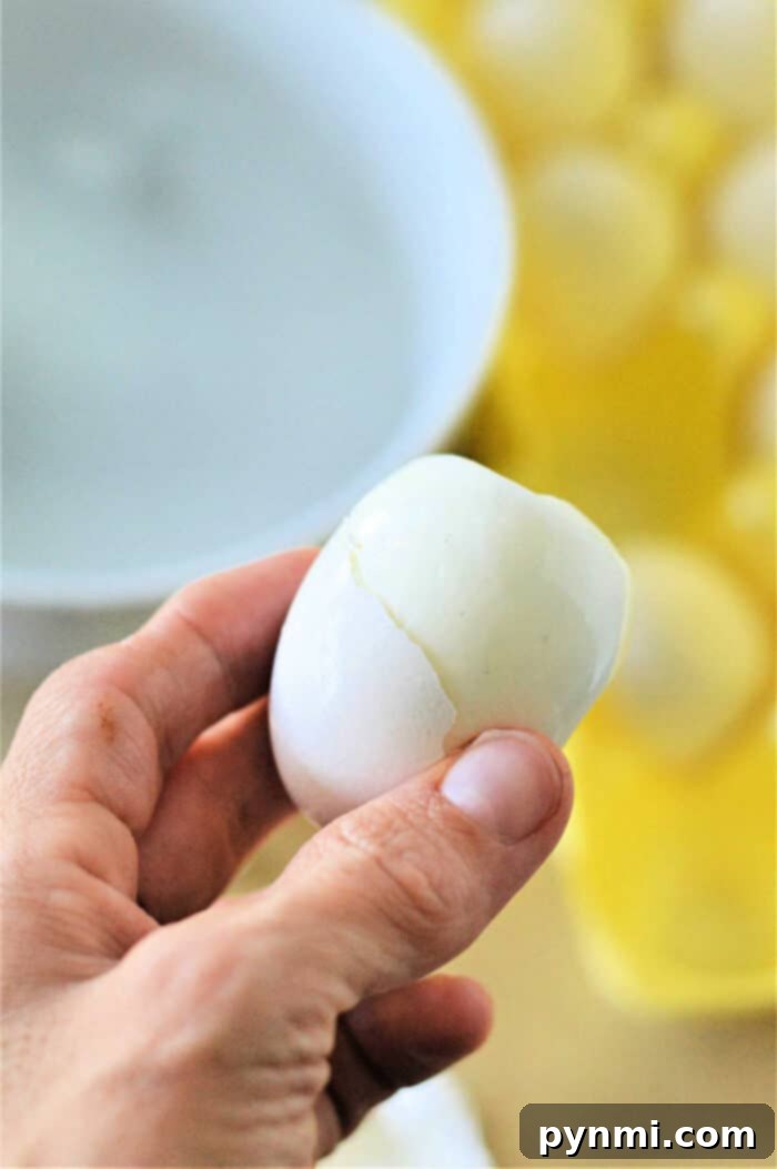 Your Toaster Oven's Secret for Perfect Hard Boiled Eggs 6 A hand holding a partially peeled boiled egg. In the background, there is a white bowl and a yellow egg carton containing several boiled eggs, hinting they might have been cooked using an eggs in toaster oven method.