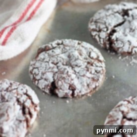 Chocolate crinkle cookies, fresh from the toaster oven, dusted with powdered sugar on a metal baking sheet near a striped cloth.