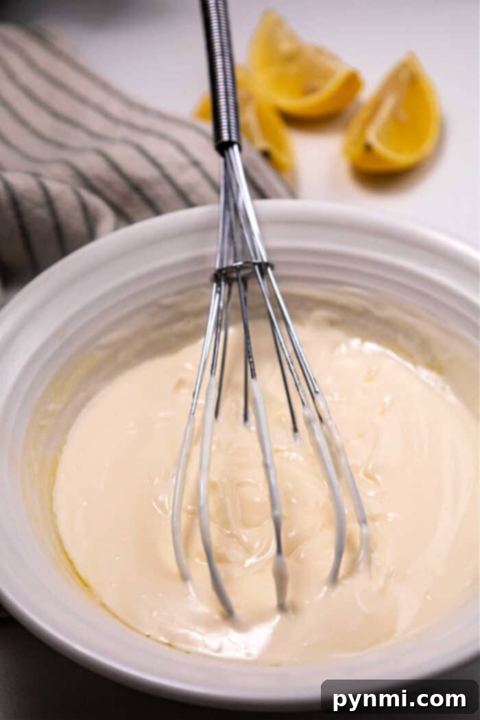 A whisk in a bowl of thick mayonnaise, with lemon wedges and a striped cloth in the background—its almost reminiscent of preparing Mexican mayonnaise.