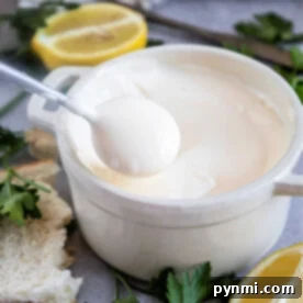A spoon dips into a white ceramic bowl of creamy Mexican mayonnaise, surrounded by lemon slices and parsley.