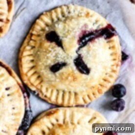 Golden-crusted air fryer blueberry hand pies, topped with sugar crystals and bursting with luscious blueberry filling, rest elegantly on a parchment-lined surface.