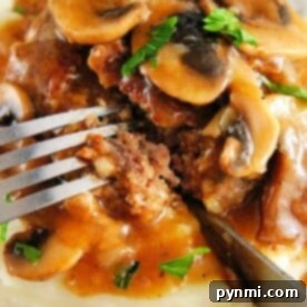 A fork cuts into a Salisbury steak with mushroom gravy atop mashed potatoes, reminiscent of delightful ground venison recipes.