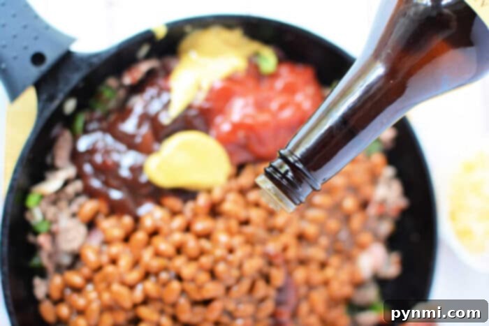 A close-up of a skillet filled with baked bean casserole, ground meat, yellow mustard, and ketchup. A bottle is seen from the top right corner, pouring additional sauce onto the mixture. The skillet handle is visible on the left side of the image.