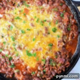 A cast iron skillet filled with baked bean casserole topped with a layer of melted cheese and garnished with chopped parsley. The dish is placed on a light wooden surface.