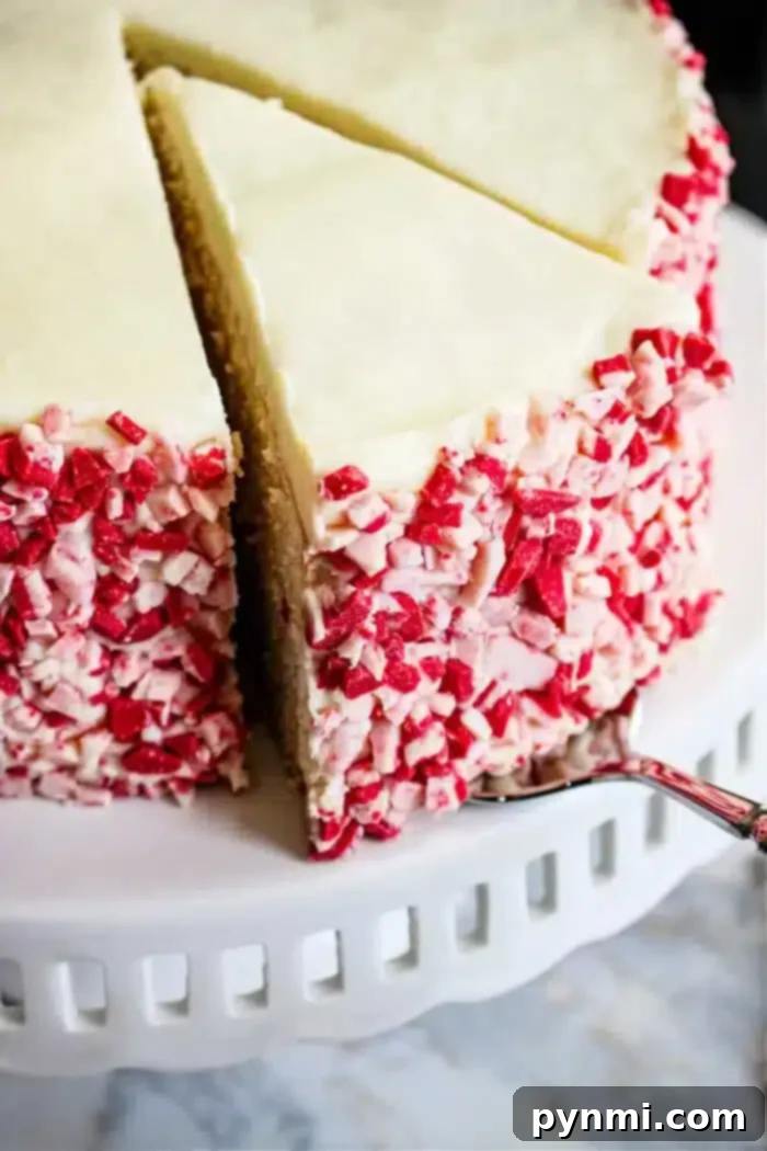 Peppermint Bliss Cake 2 A Peppermint Cake with white frosting and red and white crushed candy on the sides is displayed on a white cake stand. A slice has been cut and is being removed with a cake server.