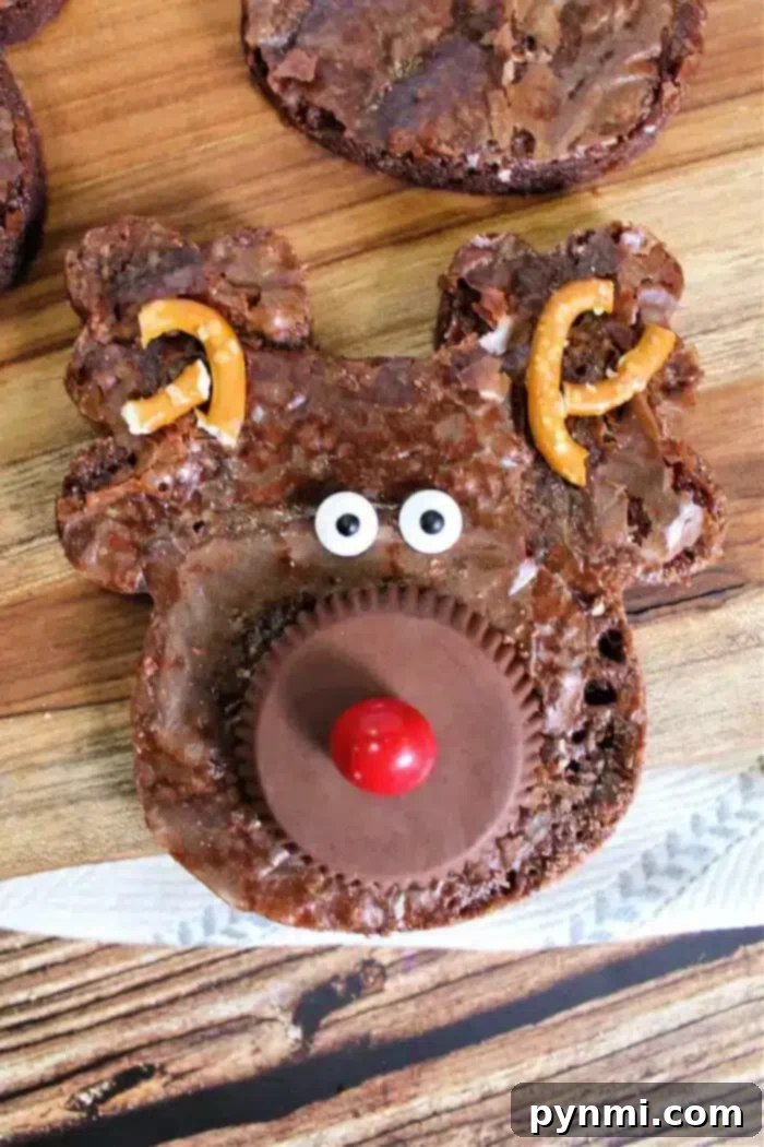 Yuletide Chocolate Squares 2 A reindeer-shaped Christmas brownie with a peanut butter cup for a nose, a red candy on top, candy eyes, and broken pretzels as antlers rests on a wooden surface. Other similar brownies are partially visible in the background.