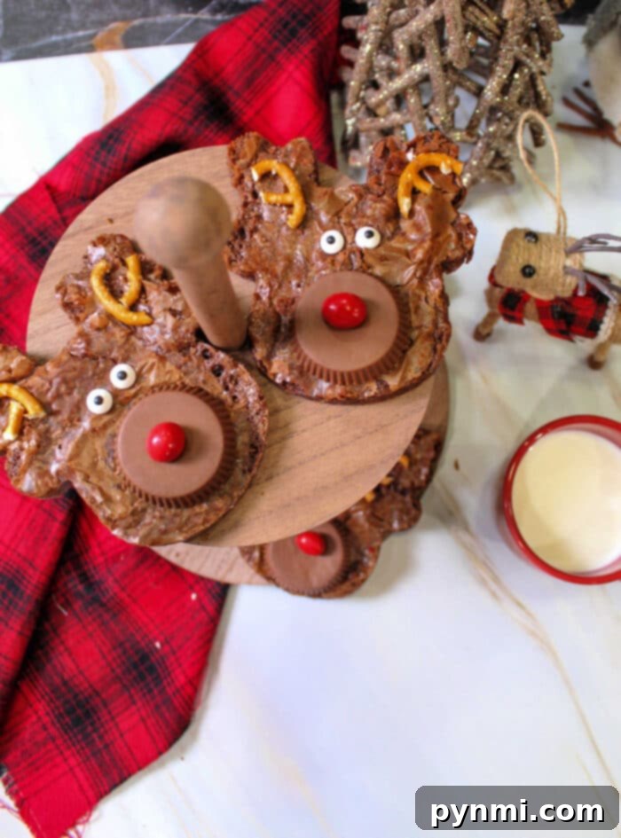 Yuletide Chocolate Squares 6 Reindeer-themed brownies with pretzel antlers and candy eyes sit alongside Christmas brownies on a wooden stand, next to a red plaid cloth and a drink.