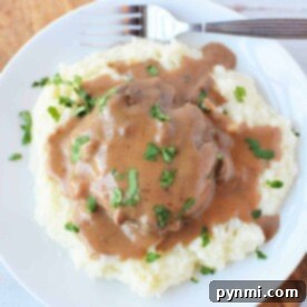 Plate of mashed potatoes topped with brown gravy and chopped herbs, served alongside tender Crockpot Salisbury Steak with a fork.