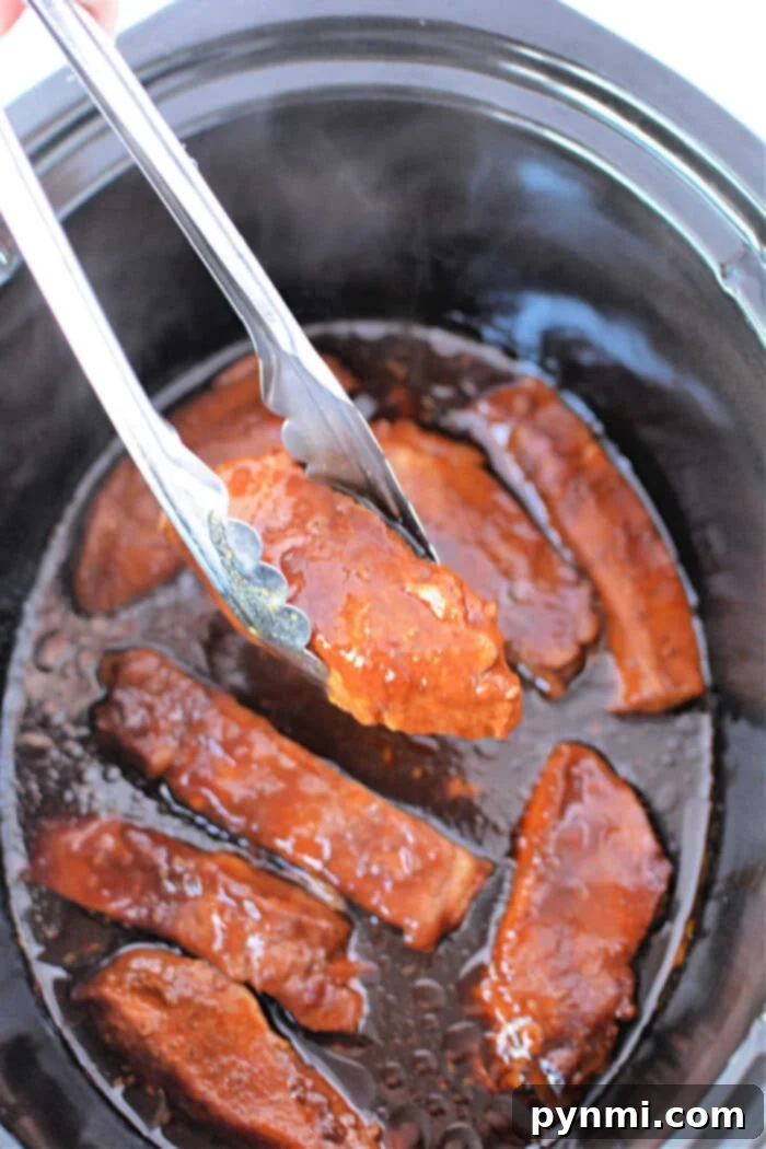 Homestyle Slow Cooker Country Ribs 6 Tongs holding saucy, cooked country ribs hover over a slow cooker brimming with country-style ribs.