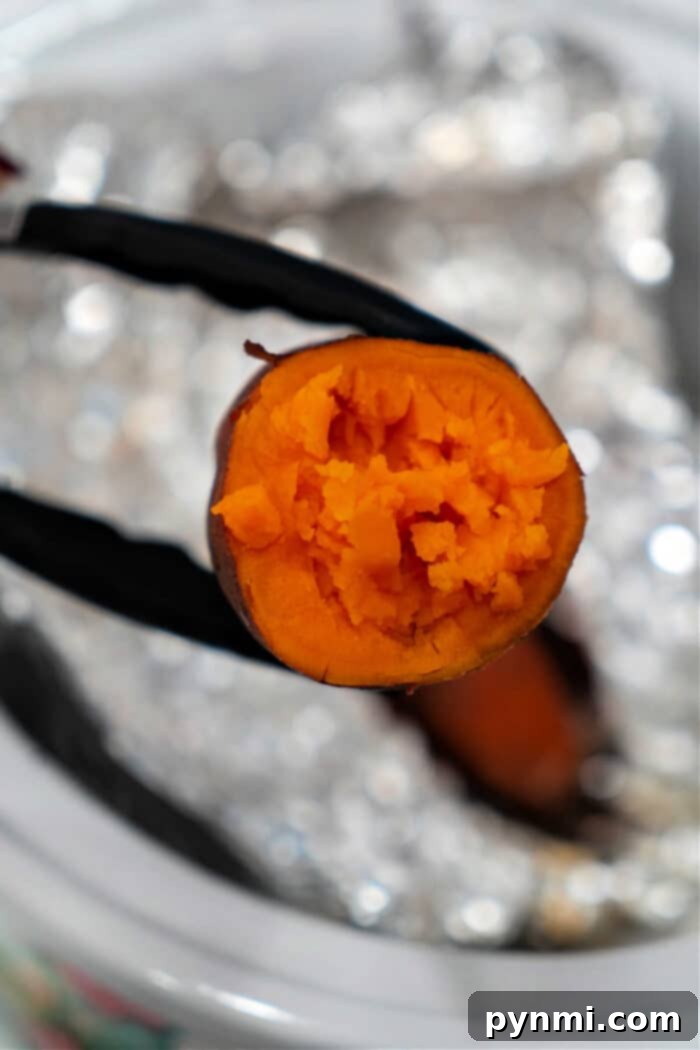 Set It and Forget It Whole Sweet Potatoes 7 A cooked sweet potato held by tongs, with vibrant orange flesh visible against a blurred kitchen backdrop, showcases the delightful simplicity of crockpot sweet potatoes.