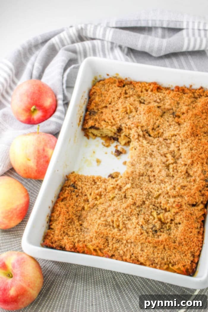 Spiced Apple Comfort Bake 6 A beautifully baked sweet apple kugel in a white dish, artfully arranged with fresh apples and a charming striped cloth nearby, inviting a taste of this comforting casserole.