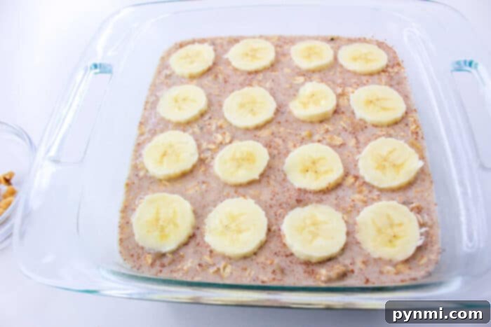 A clear glass baking dish holding the uncooked Banana Bread Baked Oatmeal batter, expertly smoothed and elegantly topped with thin, fresh banana slices and a light sprinkle of crushed nuts, patiently awaiting its transformation in the oven.