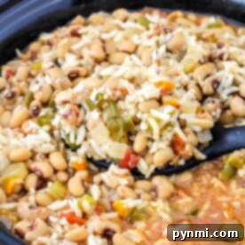 A crockpot filled with black-eyed peas, rice, vegetables, and a savory broth, with a spoon for serving.