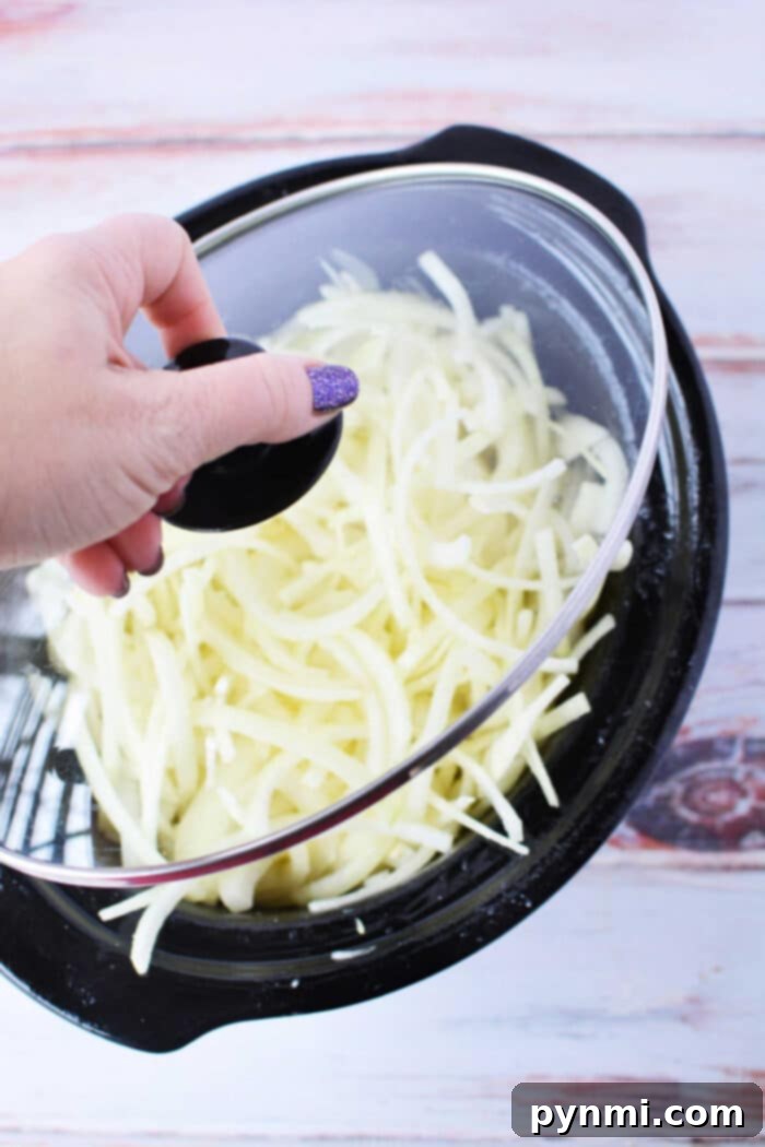 Hand lifting lid from slow cooker filled with shredded potatoes and caramelized onions.